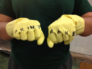 Close-up of yellow gloves with text written on fingers: PRINT MAKE