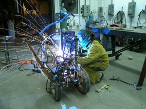 Student welding in yellow suit and face mask, sparks flying