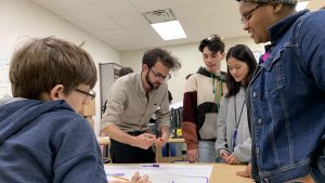 Male college student with glasses leans over table, high school students gather around