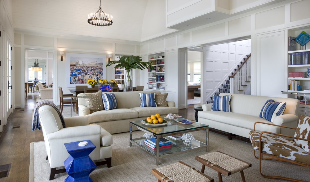 Living room with cream sofa, striped pillows, glass coffee table, textured rug, bright painting on white walls, chandelier over table